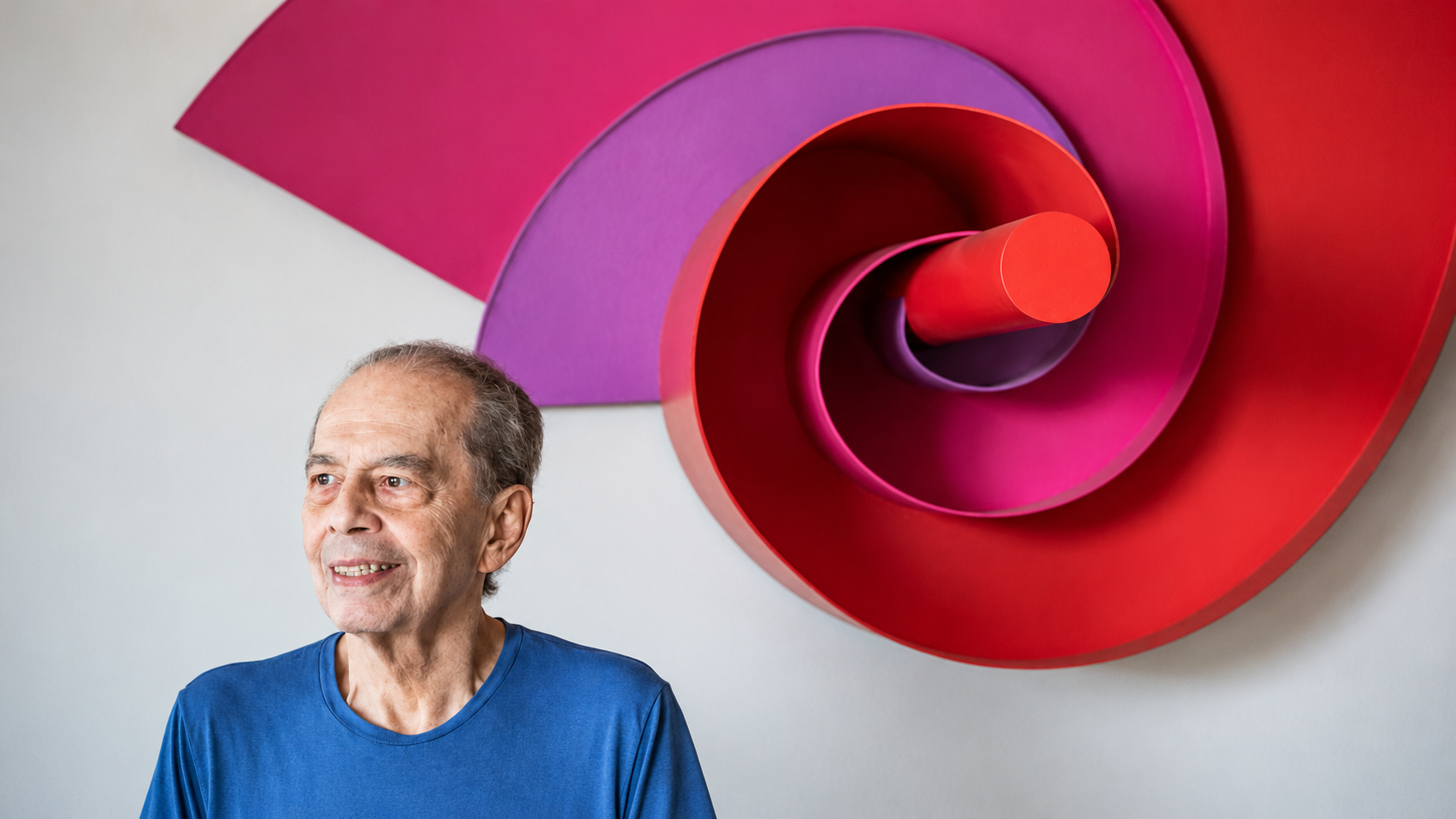 Retrato de um homem diante de uma obra geométrica tridimensional em tons de vermelho, rosa e roxo.
